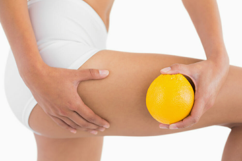 Muslos lisos de una mujer sin celulitis, junto a una naranja sostenida en la mano